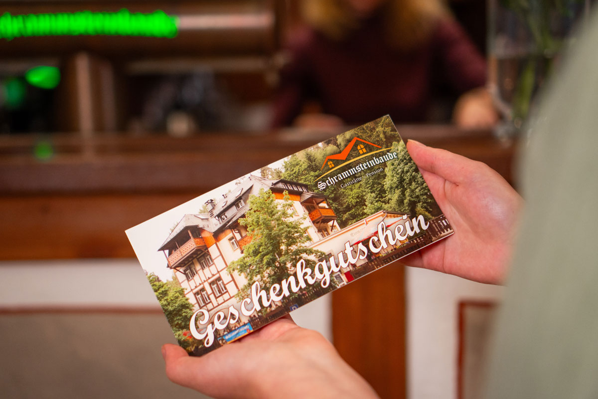 Gaststätte und Pension Schrammsteinbaude – Gutschein verschenken Hände halten einen Geschenkgutschein, der die Schrammsteinbaude in der Außenansicht und das Logo zeigt