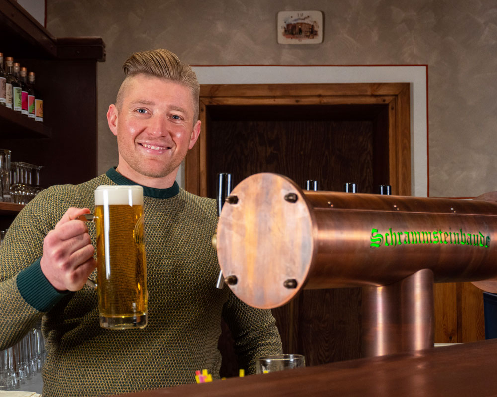Gaststube Schrammsteinbaude – Gastwirt Steve Möller Gastwirt Steve Möller steht am Tresen und hält lächelnd ein Bier in die Höhe
