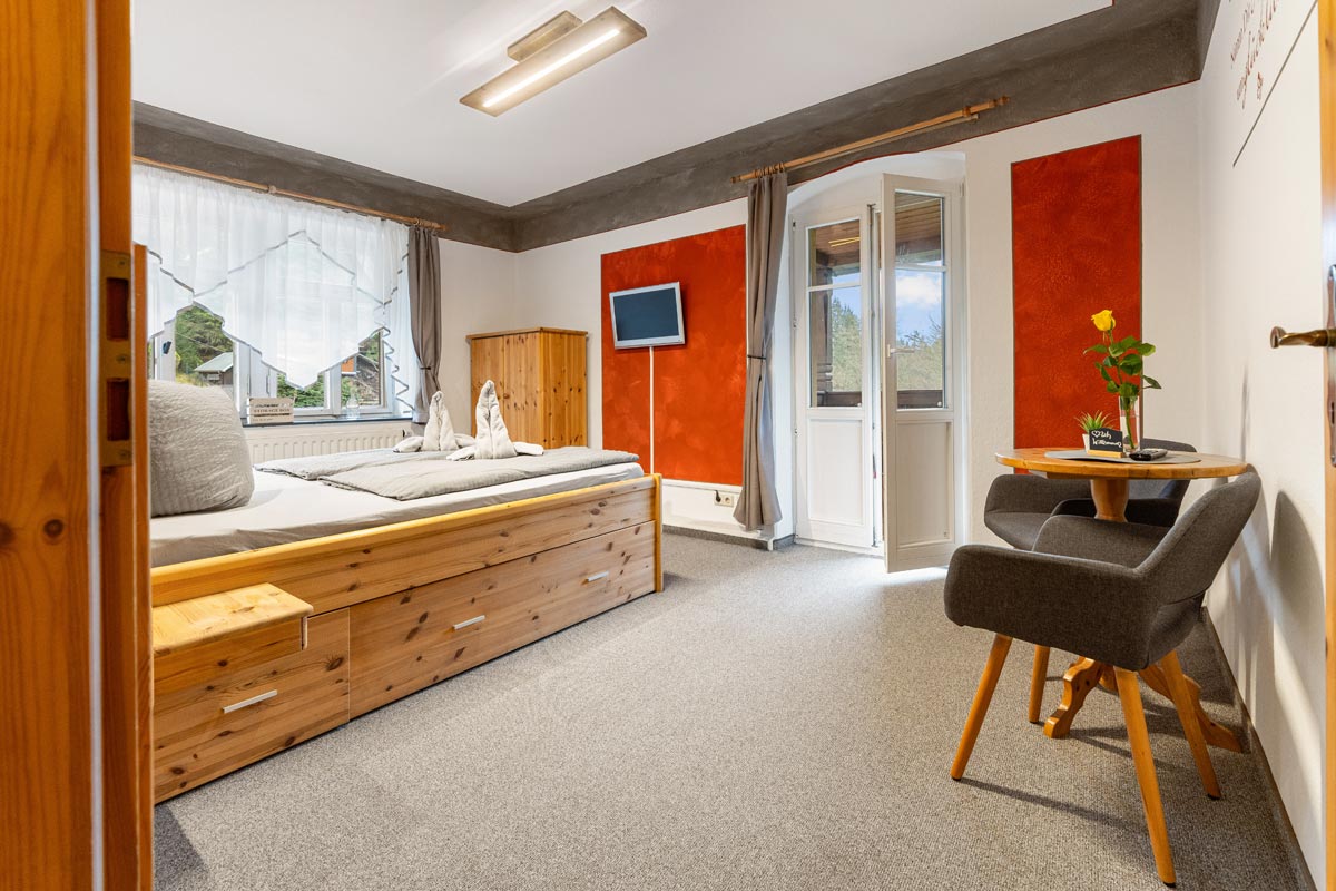Pension Schrammsteinbaude – Doppelzimmer mit Balkon – Blick auf Balkontür Ein helles Doppelzimmer mit Bett in Naturholzoptik, Sitzmöglichkeiten, Fernseher und offen stehender Balkontür.