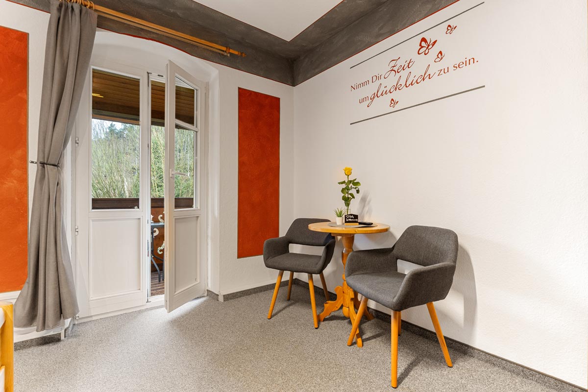 Blick in ein helles Zimmer mit Sitzgelegenheit, Tisch, Wandspruch und offen stehender Balkontür.