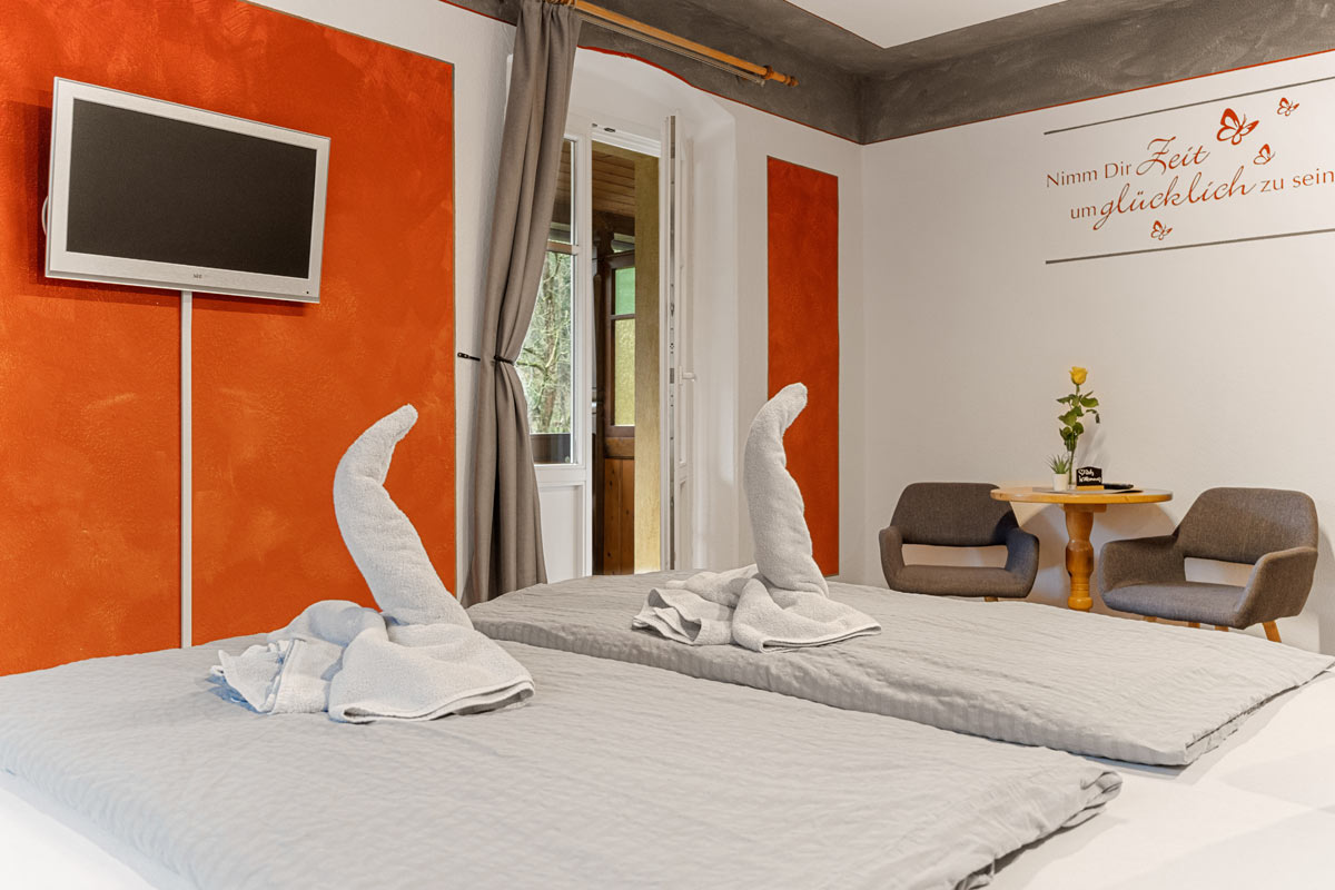 Blick in ein helles Zimmer mit Sitzgelegenheit, Tisch, Doppelbett, Flat-TV und offen stehender Balkontür.