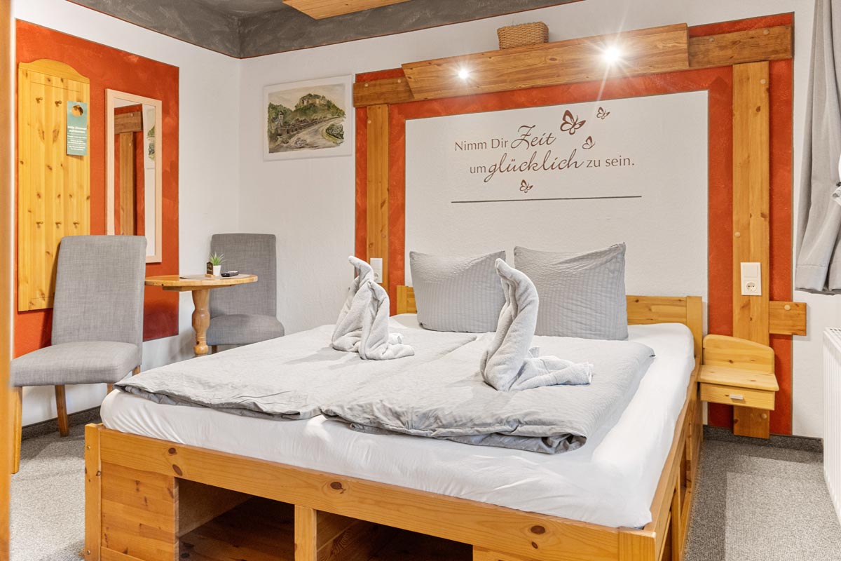 Blick in ein helles Zimmer mit Naturholzoptik, Sitzgelegenheit, Tisch, Wandspruch und Doppelbett.