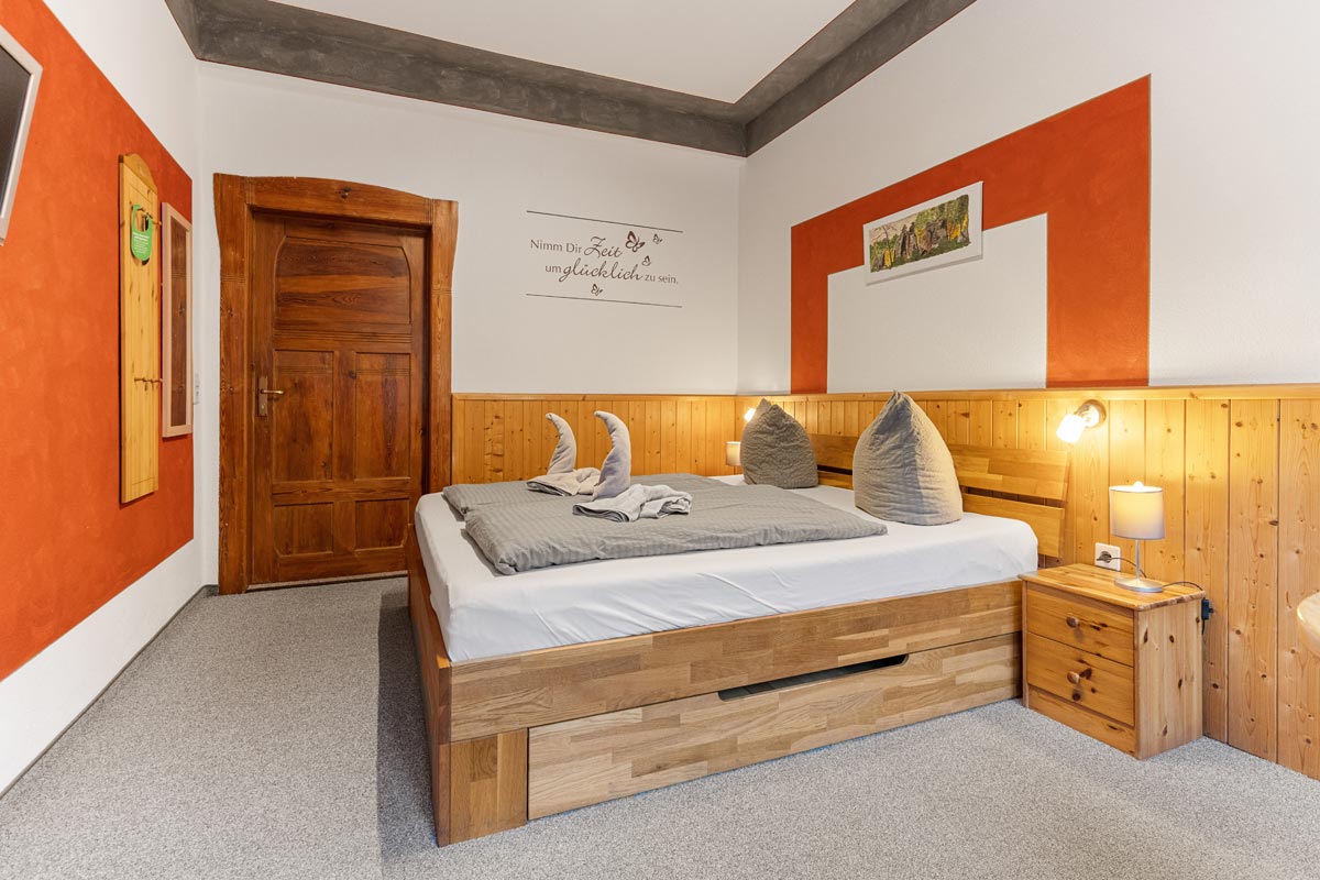 Pension Schrammsteinbaude – Doppelzimmer ohne Balkon – Blick in das Doppelzimmer Blick in ein helles Zimmer mit Naturholzoptik, Doppelbett und Wandbildern
