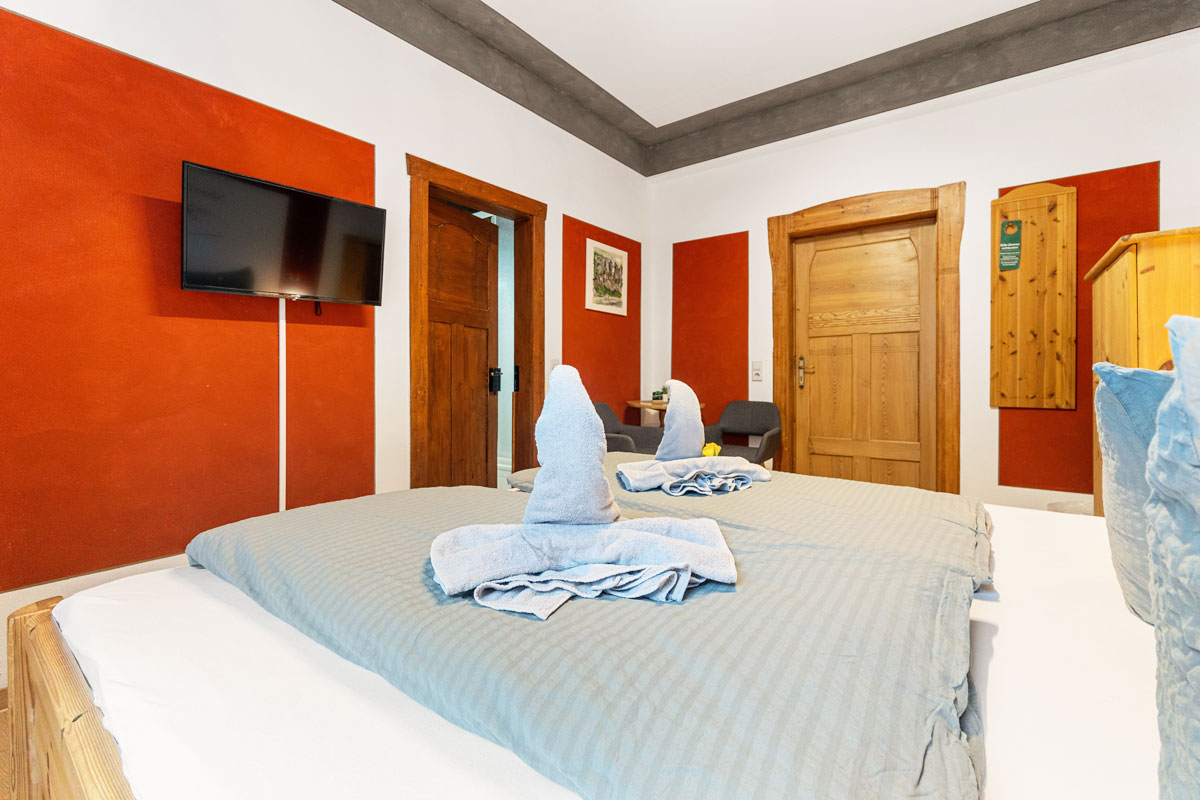 Blick in ein helles Zimmer mit Naturholzoptik, Doppelbett, Sitzgelegenheit und Flat-TV an der Wand.