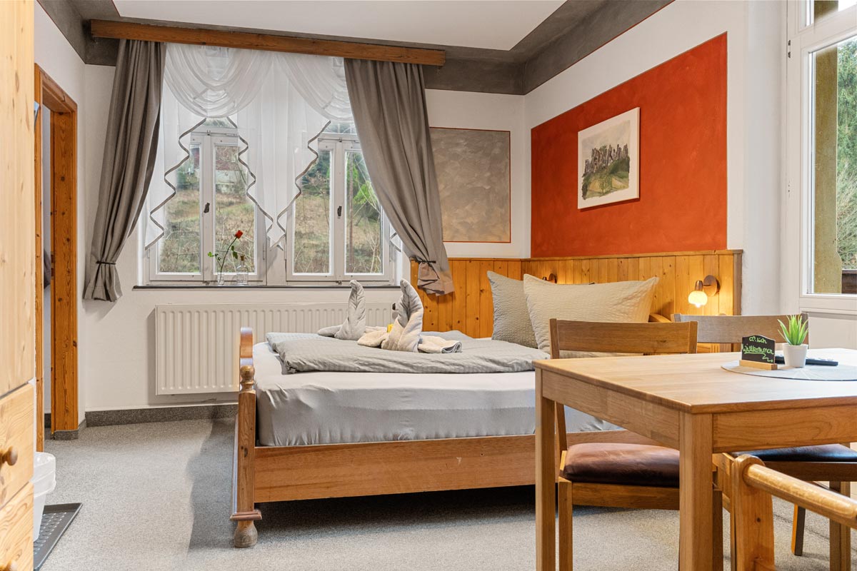 Pension Schrammsteinbaude – Familienzimmer mit Balkon – Doppelbett und Esstisch Blick in ein helles Zimmer mit Naturholzoptik, Doppelbett und Sitzgelegenheit.
