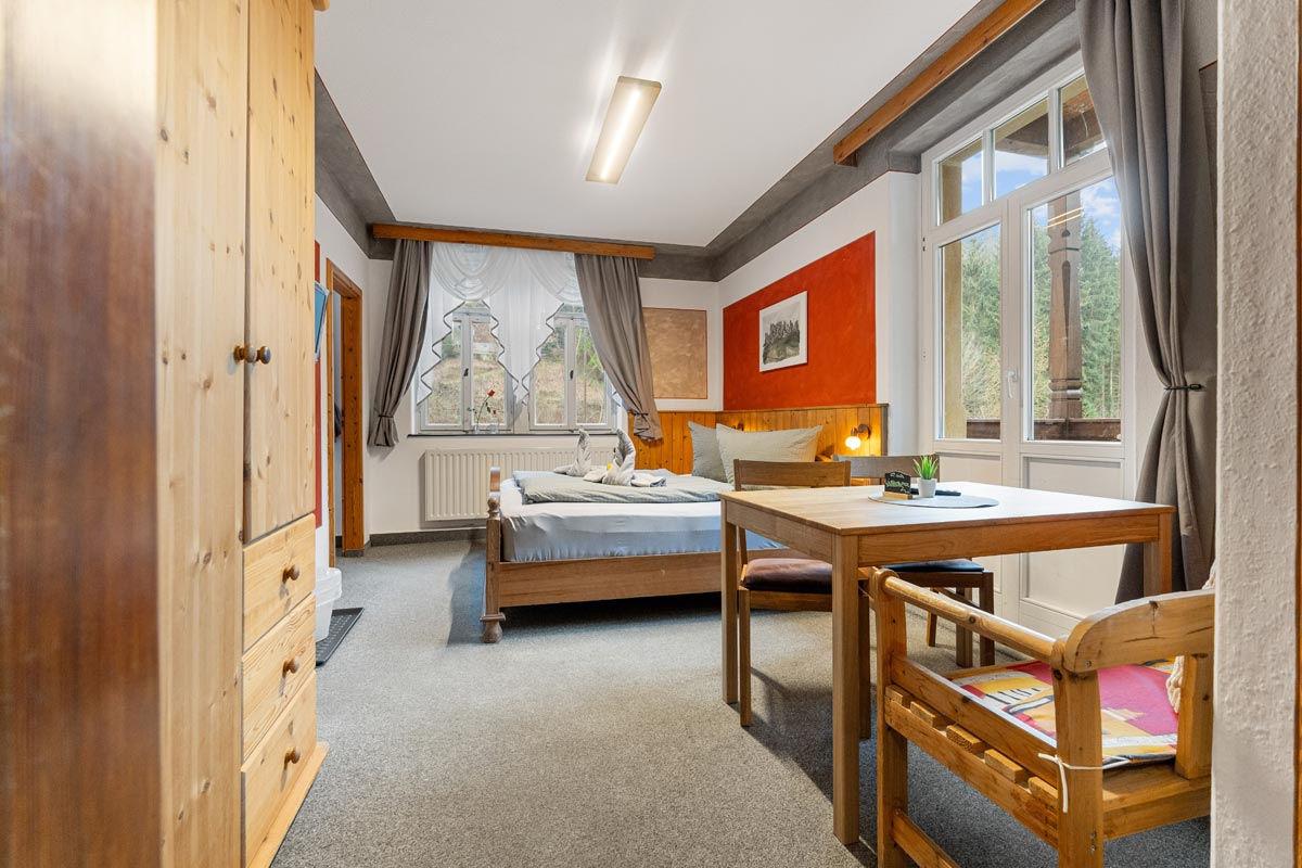 Pension Schrammsteinbaude – Familienzimmer mit Balkon – Doppelbett und Kleiderschrank Blick in ein helles Zimmer mit Naturholzoptik, Doppelbett, Sitzgelegenheit und Kleiderschrank.