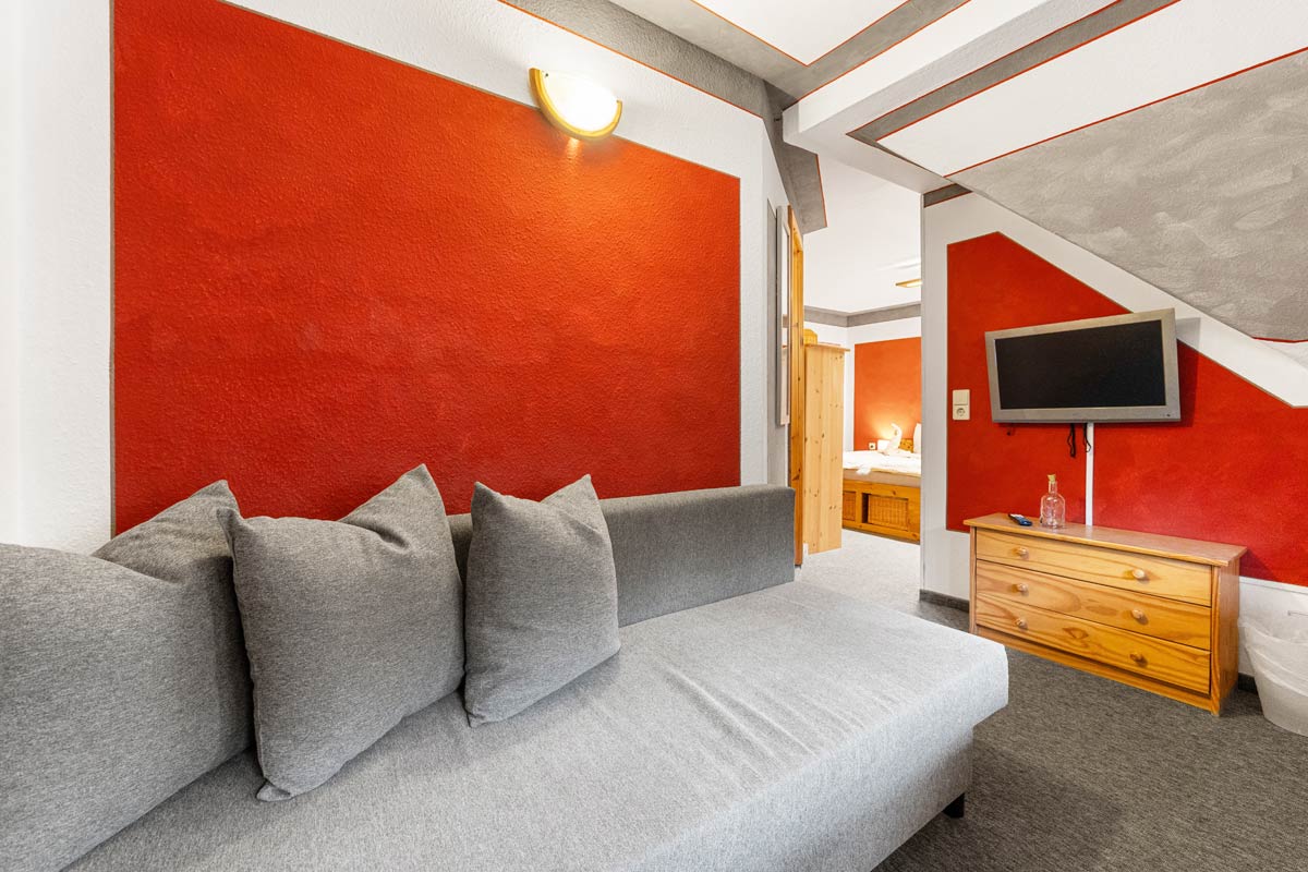 Pension Schrammsteinbaude – Familienzimmer mit Balkon – Sofa und Flat-TV Blick in ein helles Zimmer mit Naturholzoptik, Sofa und Flat-TV an der Wand.