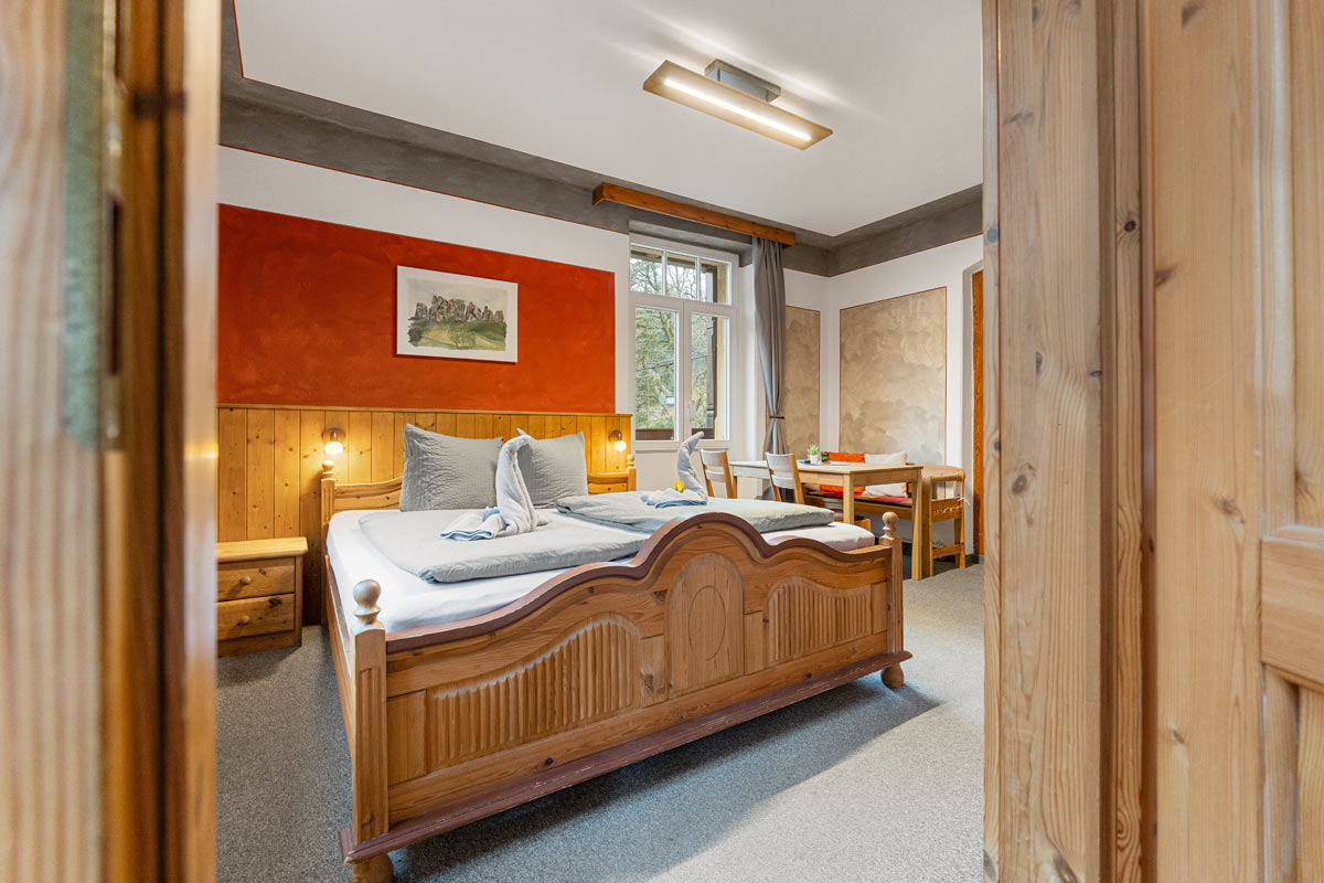 Pension Schrammsteinbaude – Familienzimmer mit Balkon und Esstisch Blick in ein helles Zimmer mit Naturholzoptik, Doppelbett, Sitzgelegenheit und Wandbild.