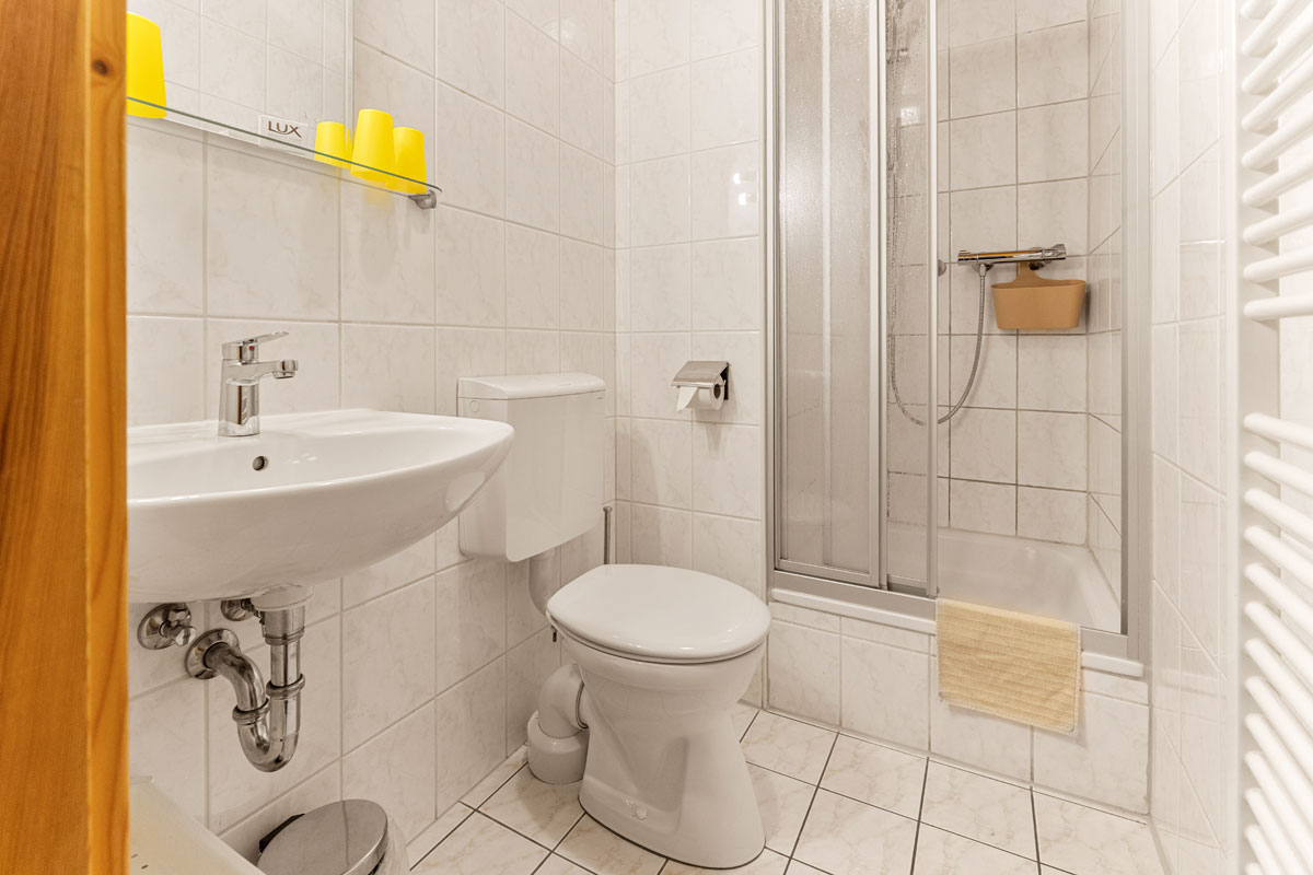 Pension Schrammsteinbaude – Familienzimmer ohne Balkon – Badezimmer mit WC und Dusche Blick in ein fensterloses Badezimmer mit weißen Fliesen, Dusche, WC und Waschbecken.