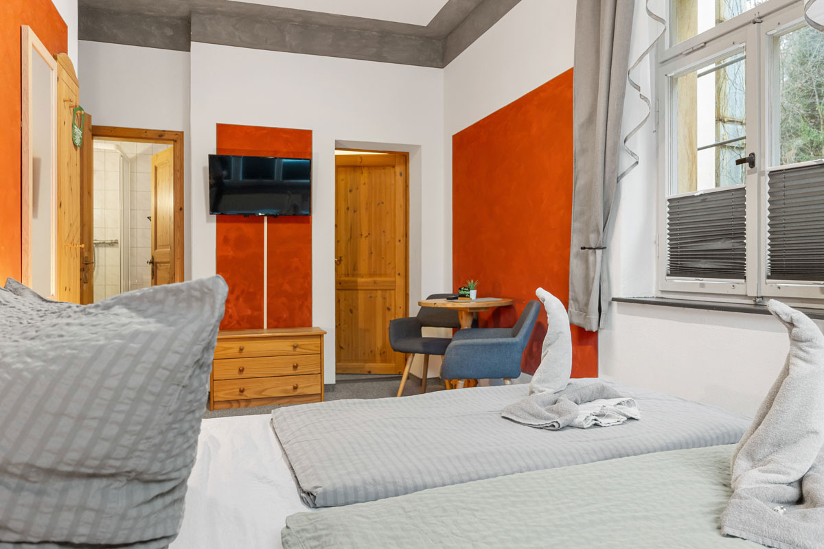 Blick in ein helles Zimmer mit Naturholzoptik, Doppelbett, Sitzgelegenheit und Flat-TV an der Wand.