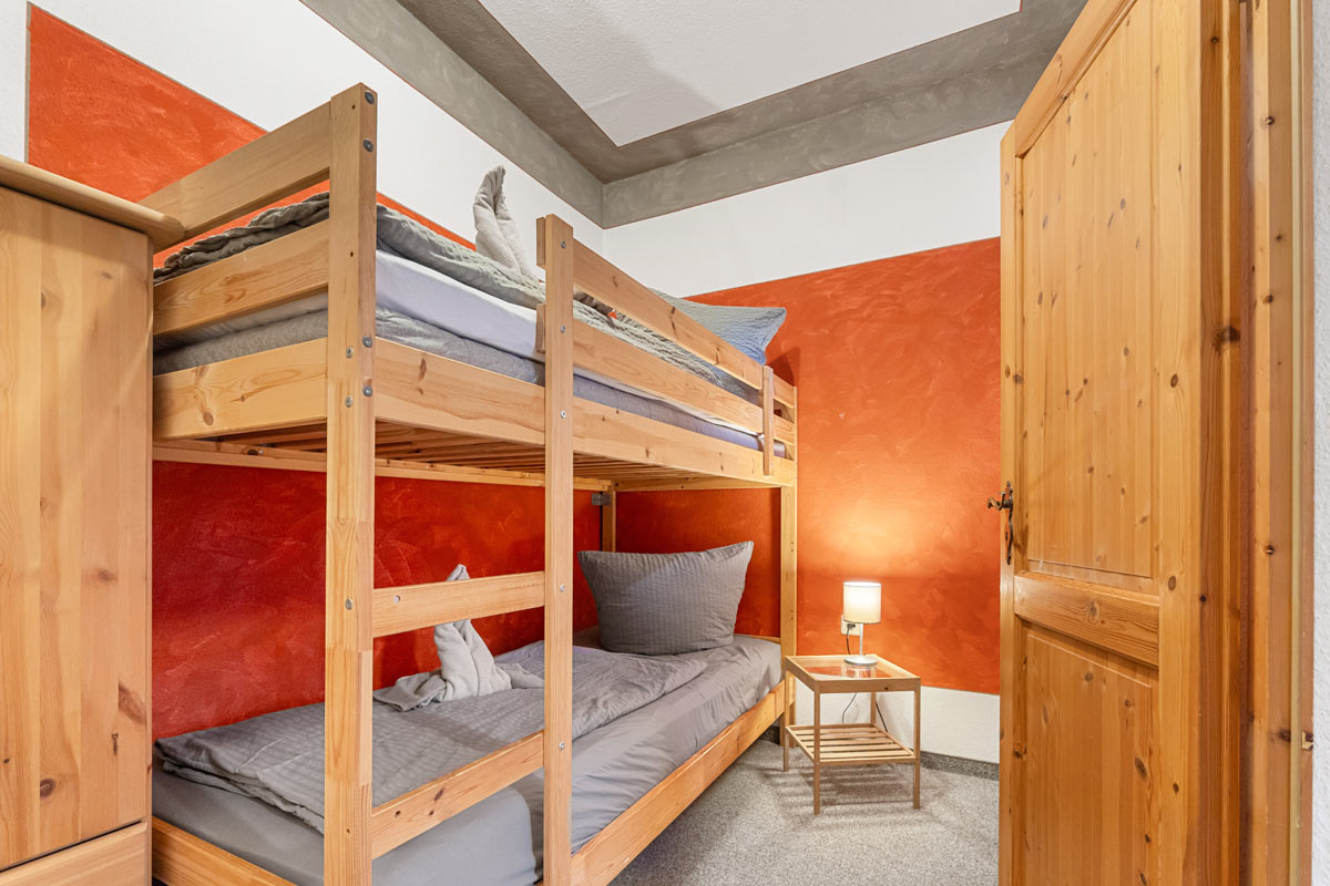 Pension Schrammsteinbaude – Familienzimmer ohne Balkon mit Stockbett Blick in ein helles Zimmer mit Naturholzoptik, Stockbett und Nachttisch mit Lampe.