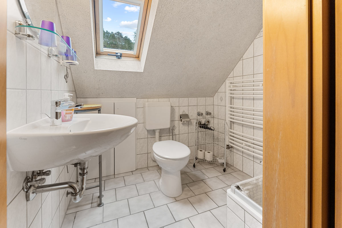 Blick in ein weiß gefliestes Badezimmer im Dachgeschoss mit Fenster, Dusche, WC und Waschbecken.