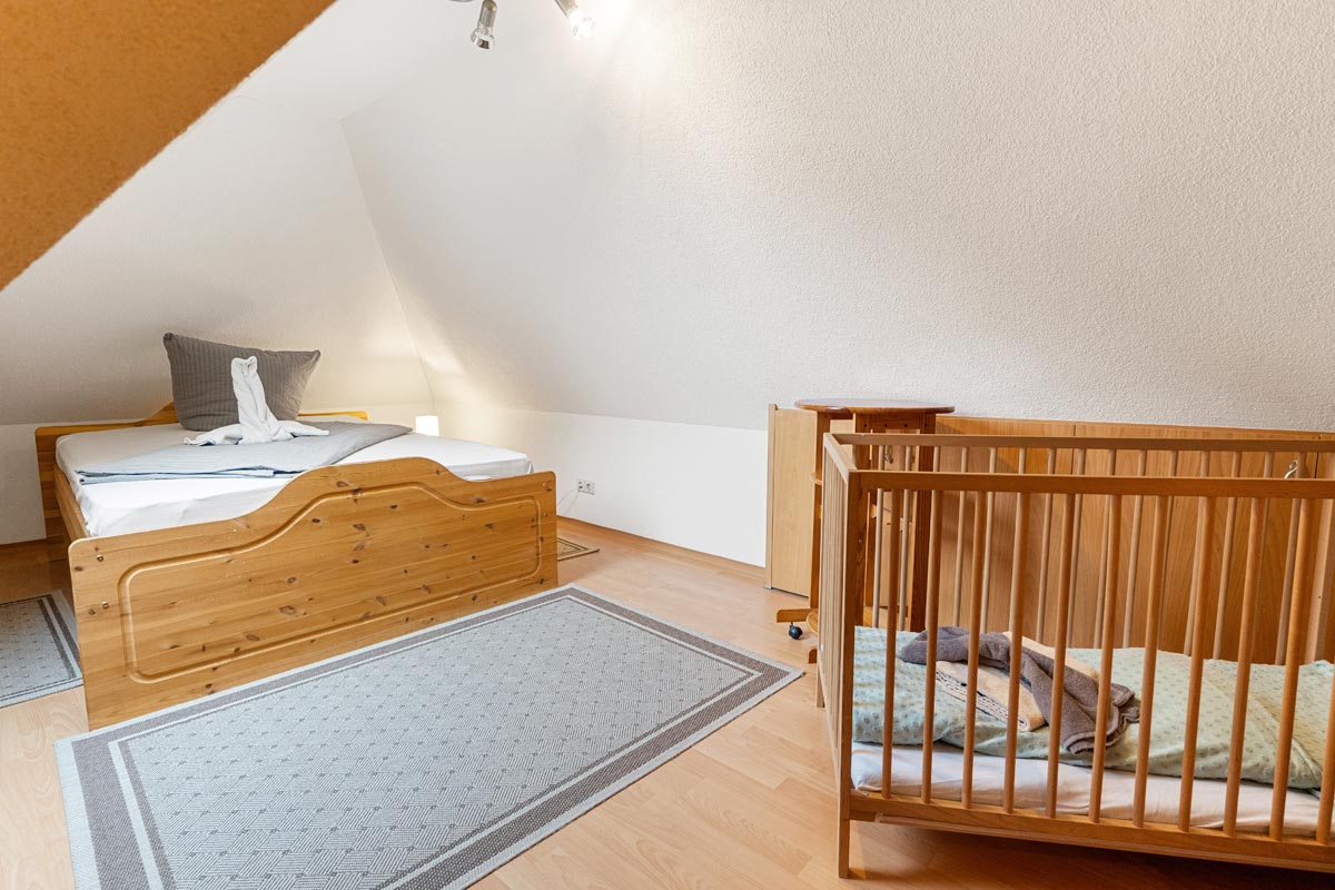 Pension Schrammsteinbaude – Ferienwohnung im Dachgeschoss – Schlafzimmer mit Kinderbett Blick in ein helles Zimmer im Dachgeschoss mit Naturholzoptik, Doppelbett und Kinderbett.