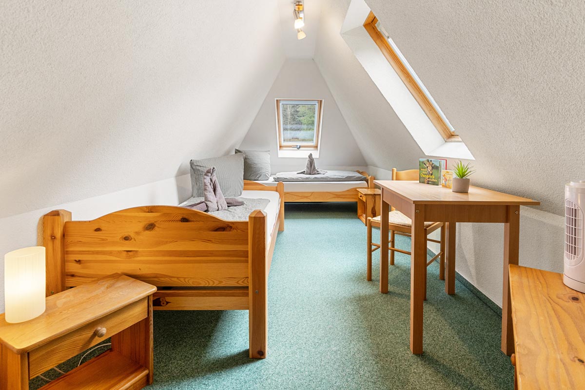 Pension Schrammsteinbaude – Ferienwohnung im Dachgeschoss – Schlafzimmer mit Einzelbetten Blick in ein helles Zimmer im Dachgeschoss mit Naturholzoptik, Einzelbetten und Sitzgelegenheit.