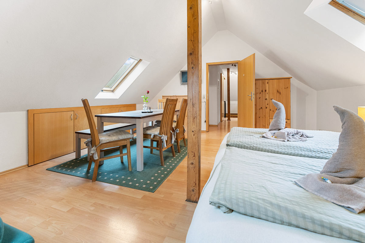 Pension Schrammsteinbaude – Ferienwohnung im Dachgeschoss – Schlafgelegenheit und Esstisch Blick in ein helles Zimmer im Dachgeschoss mit Naturholzoptik, Schlafgelegenheit und Esstisch.
