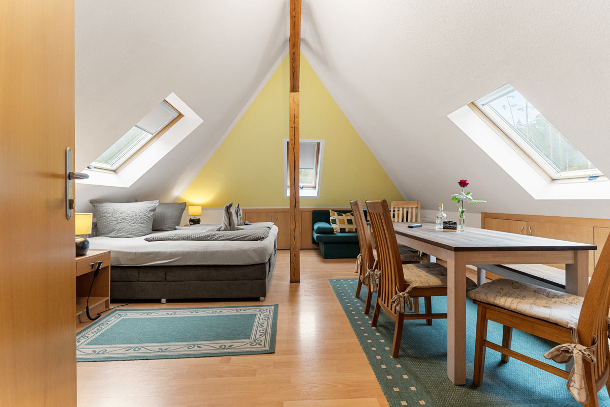 Blick in ein helles Zimmer im Dachgeschoss mit Naturholzoptik, Schlafgelegenheit, Sofa und Esstisch.
