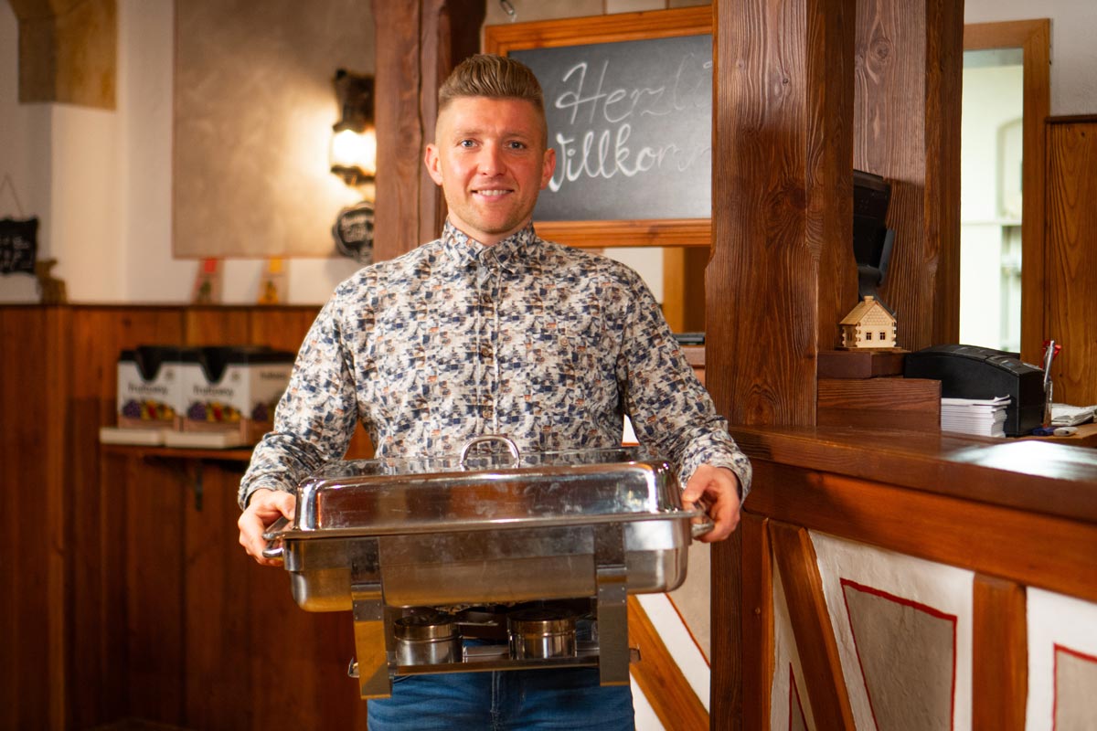 Schrammsteinbaude – Catering- und Eventservice Gastwirt Steve Möller steht vor der Rezeption und trägt einen Speisenwärmer